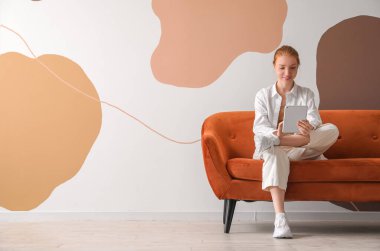 Duvarın yanındaki kırmızı koltukta tablet bilgisayar kullanan güzel bir kadın.