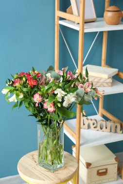 Mavi duvarın yanındaki tabure ve raf ünitesinde güzel alstromerya çiçekleri olan vazo.