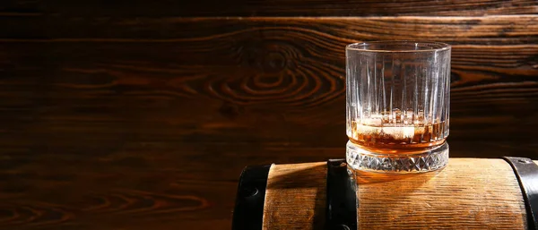 Glass of whiskey on barrel against wooden background with space for text