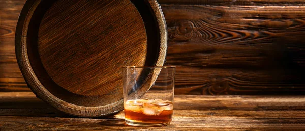 Glass of whiskey and barrel on wooden background with space for text