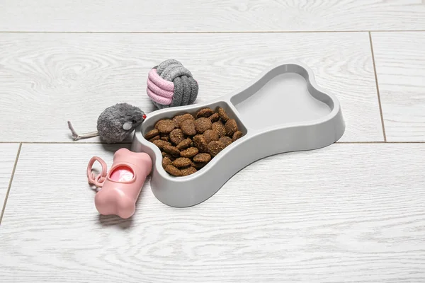 Bowl of dry pet food, waste bags and toys on light wooden background