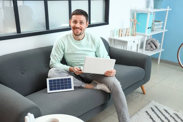 stock image Handsome man charging laptop with portable solar panel on sofa at home