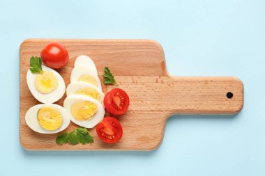 Board with delicious boiled eggs, tomatoes and parsley on blue background