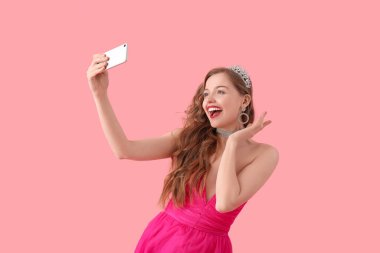 Young woman in tiara and bright prom dress taking selfie on pink background