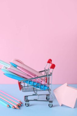 Farklı kalemleri, kalemleri ve renk arkaplanı üzerine yapışkan notları olan alışveriş arabası.