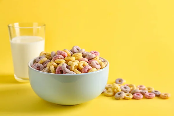 Bowl of colorful cereal rings and glass of milk on yellow background