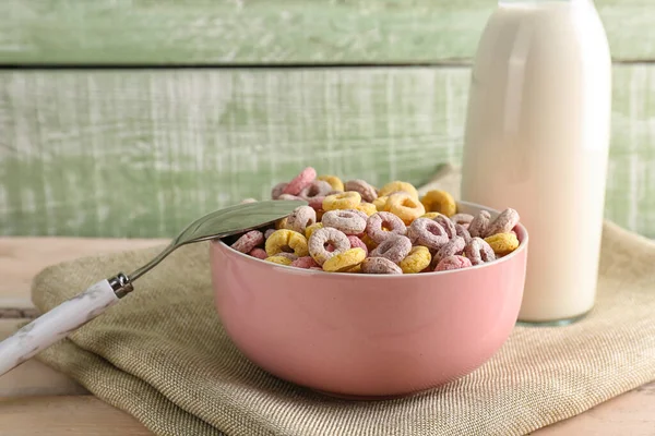 Bowl of colorful cereal rings and bottle with milk on white wooden table near green wall