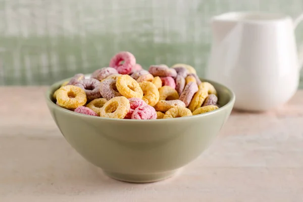Bowl of colorful cereal rings and pitcher with milk on white wooden table near green wall