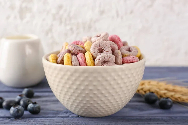 Bowl of colorful cereal rings, blueberries and wheat ears on blue wooden table near white wall