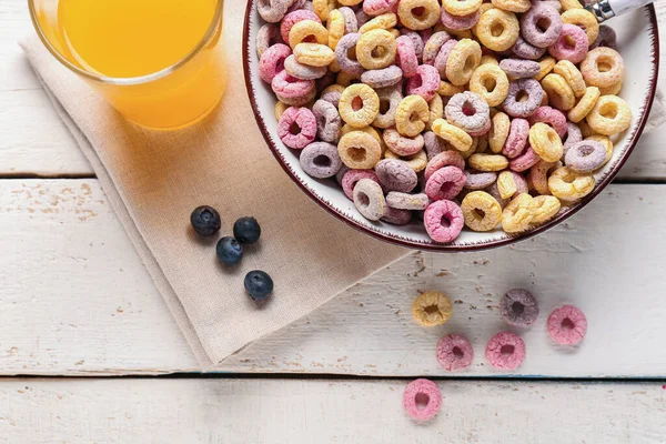 Bowl with colorful cereal rings, glass of juice and blueberries on white wooden background