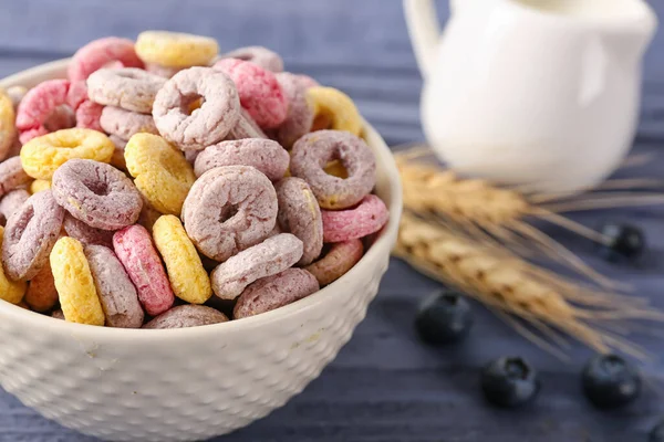 Bowl of colorful cereal rings, wheat ears and blueberries on blue wooden background