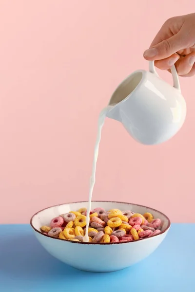 雌性手将牛奶从水罐倒入碗中 将五彩缤纷的麦片环放在粉色背景的蓝色桌子上 — 图库照片