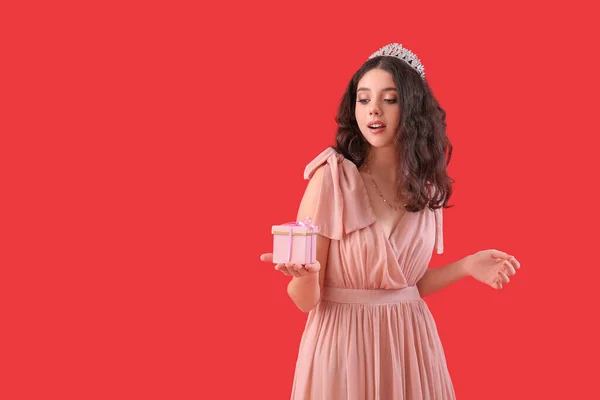Teenage girl in tiara and prom dress with gift on red background