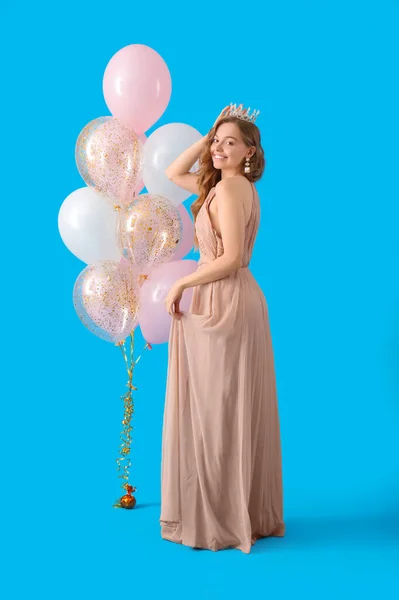 Young woman in crown and prom dress with balloons on blue background