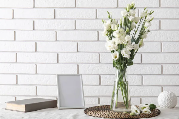 Vase with beautiful eustoma flowers, book and blank picture frame on table near light brick wall