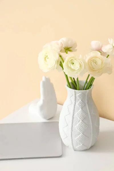 Vase with ranunculus flowers and laptop on table near beige wall, closeup