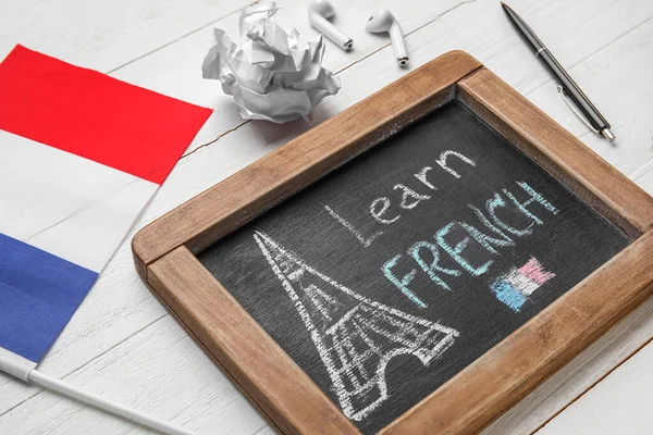 Chalkboard with text LEARN FRENCH, flag, crumpled paper and earphones on white wooden background, closeup