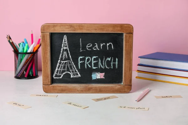 Chalkboard with text LEARN FRENCH, stationery and books on table near pink wall