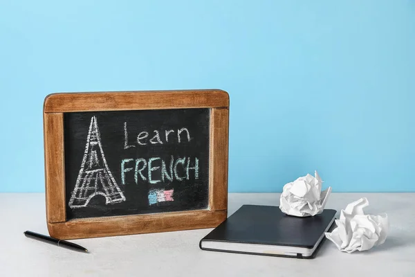 Chalkboard with text LEARN FRENCH, notebook and crumpled paper on table near blue wall