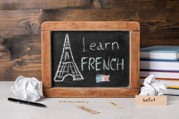 Chalkboard with text LEARN FRENCH, crumpled paper and books on table near dark wooden wall