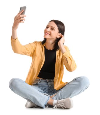 Pretty young woman with smartphone taking selfie against white background