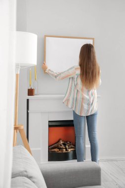 Young woman hanging blank frame on light wall at home, back view