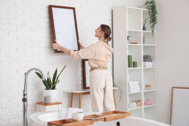Young woman hanging blank frame on white brick wall in bathroom