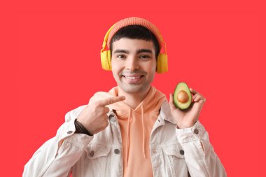 Kulaklıklı genç adam kırmızı arka planda avokadoyu işaret ediyor.