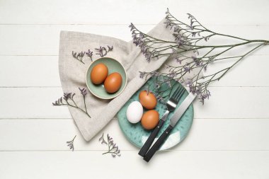 Beyaz ahşap arka planda Paskalya yumurtaları, çatal bıçak takımı ve çiçek dekoru.
