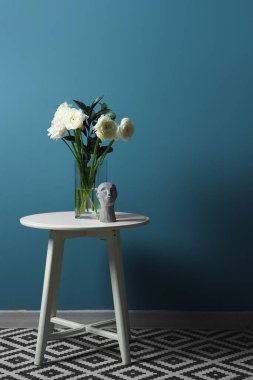 Mavi duvarın yanındaki masanın üzerinde ranunculus çiçekleri ve dekor olan vazo.