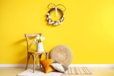 Chair with flowers in vase, pillows, pouf and Easter wreath on yellow wall