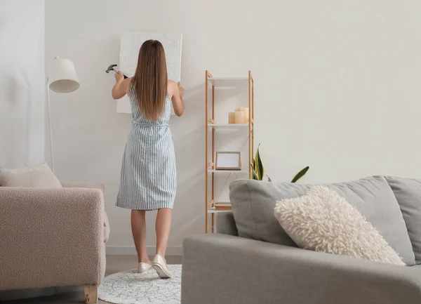 Young woman with hammer hanging painting on light wall at home, back view