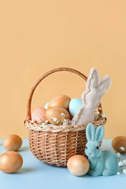 İçinde paskalya yumurtaları, tavşanlar ve bej arka planda mavi masada çigila çiçekleri olan sepet.