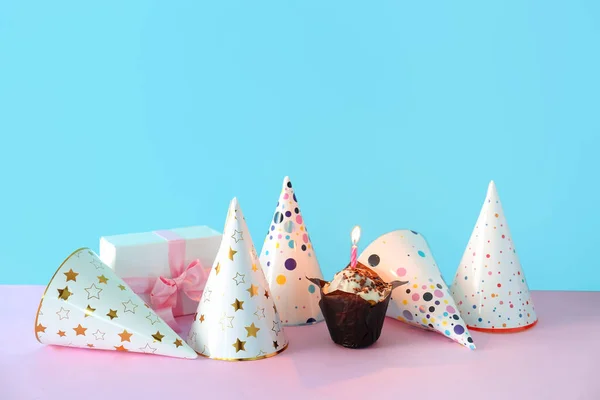Birthday cupcake, party hats and gift box on color background