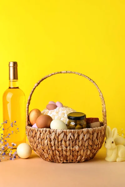 Basket with Easter eggs, cake and bottle of wine on beige table against yellow background
