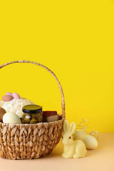 Basket with Easter eggs, cake and bunny on beige table against yellow background