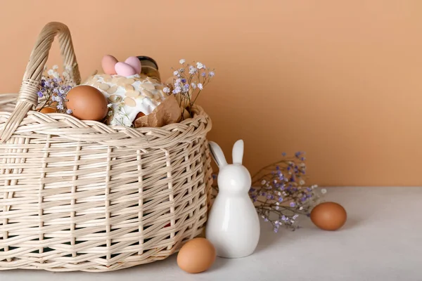 Basket with Easter eggs, cake, gypsophila flowers and bunny on white table near against beige background
