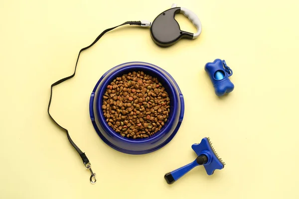 Bowl of dry pet food, grooming brush, leash and waste bags on color background