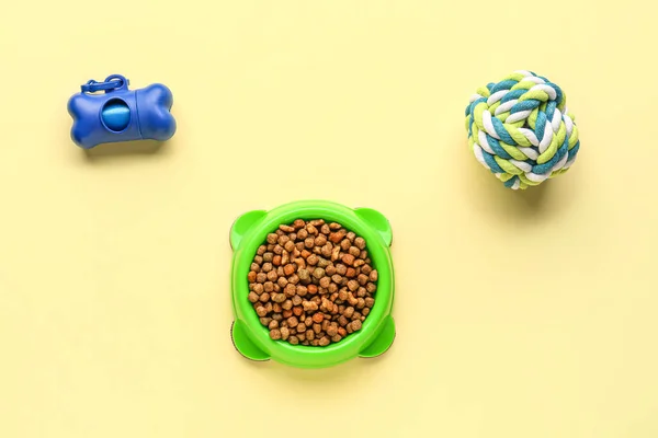 Bowl with dry pet food, toy and waste bags on color background