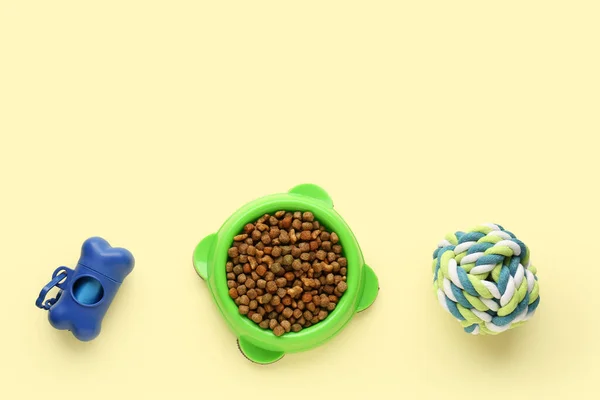 Bowl with dry pet food, toy and waste bags on color background