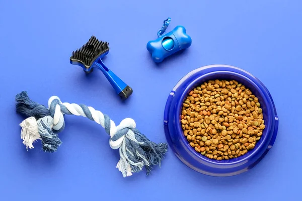 Bowl of dry pet food, toy, waste bags and grooming brush on color background