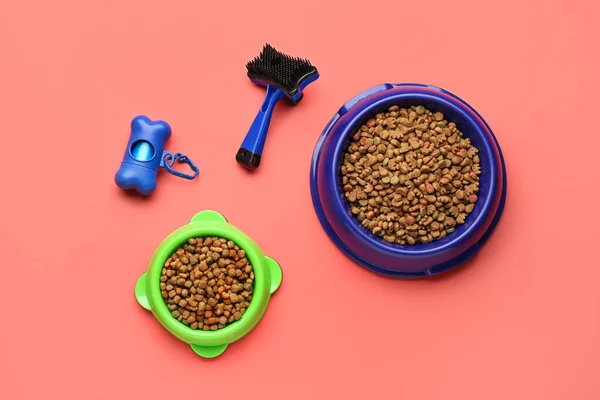 Bowls of dry pet food, grooming brush and waste bags on color background