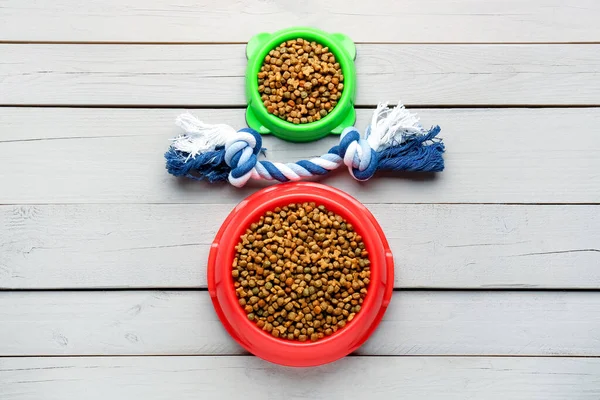 Bowls of dry pet food and toy on light wooden background