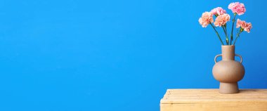 Mavi duvarın yanındaki tahta kutunun üzerinde güzel karanfil çiçekleri olan vazo. Tasarım için pankart