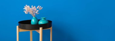 Mavi duvarın yanında vazoları ve çiçek dekoru olan şık bir masa. Tasarım için pankart