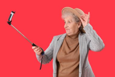 Cep telefonlu yaşlı bir kadın kırmızı arka planda selfie çekiyor.