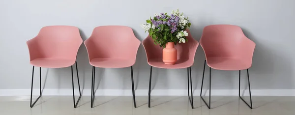 Pink armchairs with beautiful lilac flowers in vase near light wall in room