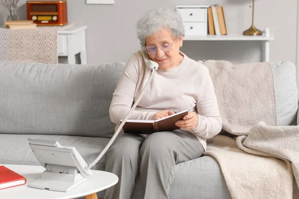 Senior woman with notebook talking by telephone at home