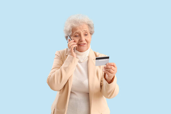 Senior woman with credit card talking by mobile phone on blue background