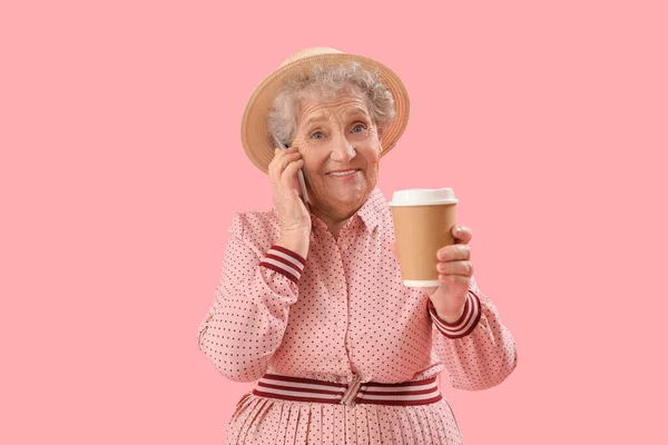 Senior woman with cup of coffee talking by mobile phone on pink background